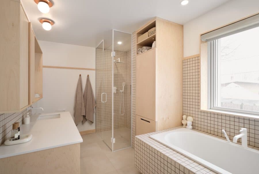Salle de bain minimaliste en bois clair avec douche vitrée, baignoire blanche et carrelage quadrillé.
