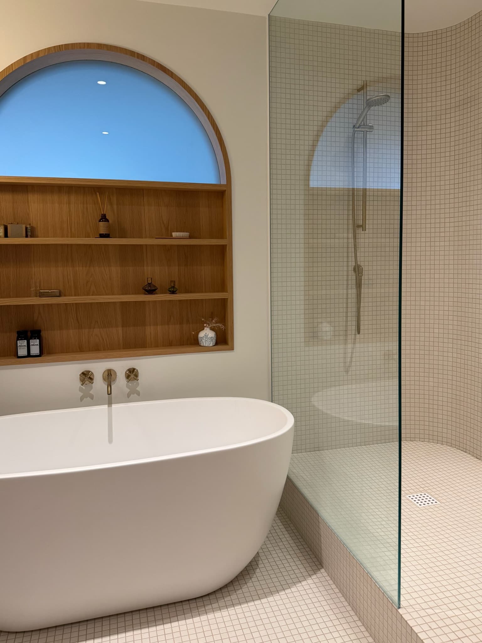 Salle de bain moderne avec baignoire blanche, étagères en bois, fenêtre arquée et douche vitrée.