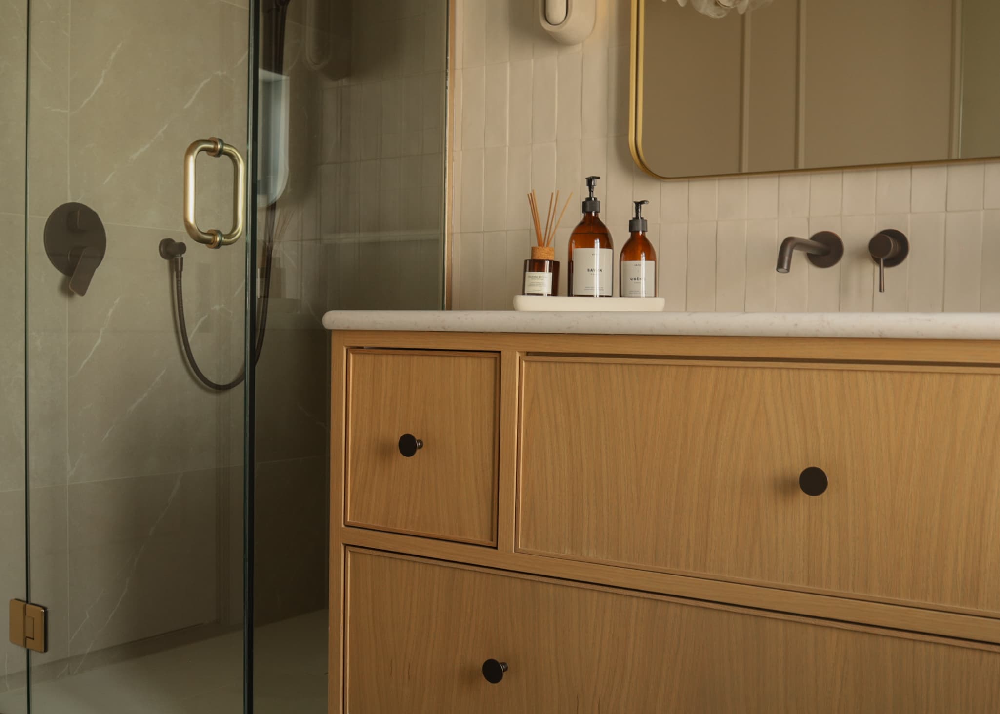 Meuble de salle de bain en bois, comptoir en marbre, flacons ambrés et douche vitrée.
